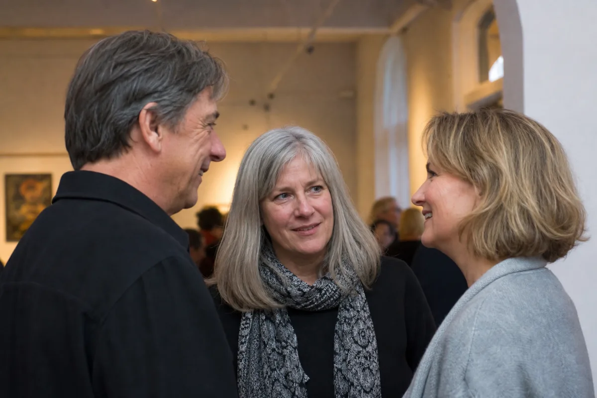 Three mature adults in their 50s and 60s talking naturally at a social arts gathering